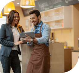 A woman in a grey suit and a man with a brown apron smile down at a tablet inside a store