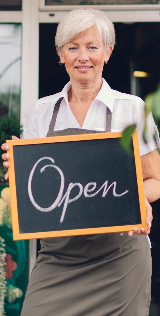 Outside of a store, an older woman in a white blouse and brown apron holds a black chalkboard with "open" written on it in cursive