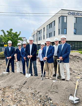 6 men and 2 women in blue blazers and white construction hats holding shovels and standing on a pile of dirt outside of Chelsea Groton Bank