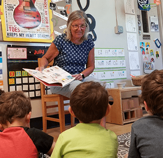An elderly woman reads a picture book to three pre-school children sitting on a classroom rug