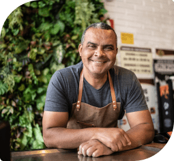 A smiling man with a grey shirt and brown apron leans on a wooden counter