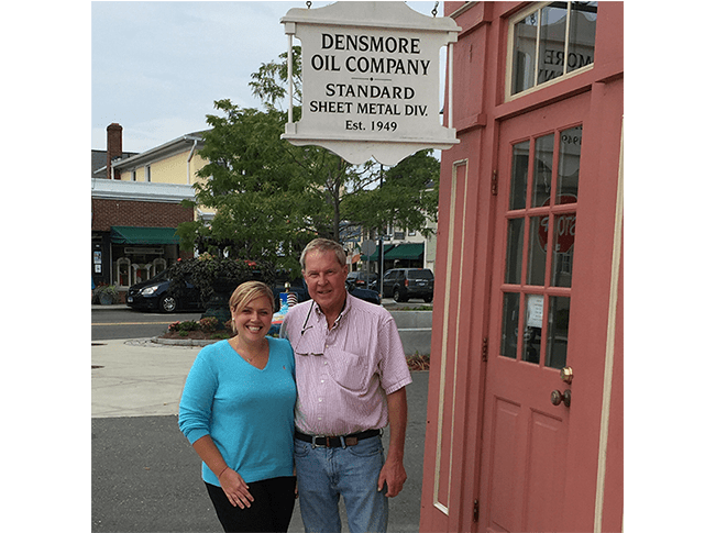 A smiling white couple standing beneath a white sign with black text that reads "Densmore Oil Company Standard Sheet Metal Div. Est. 1949"