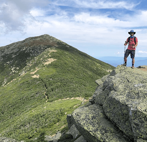 Matthew Benoit smiling with hiking sticks on top of a green mountain and bright blue sky