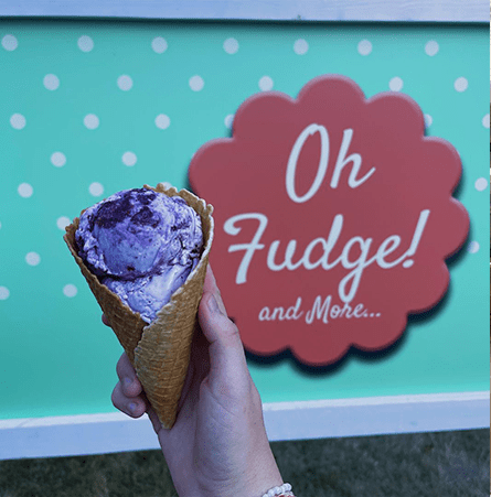 A women's hand holding purple ice cream in a waffle cone outside of a teal and red "Oh Fudge! And More..." sign