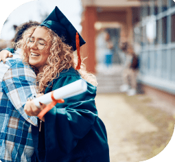 A smiling graduate in cap and gown holds diploma and hugs a loved one