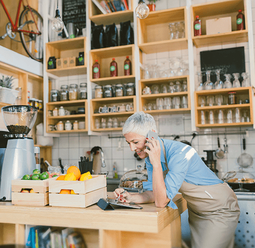 An older woman on the phone wearing a blue shirt and tan apron leans on a kitchen counter and uses a tablet