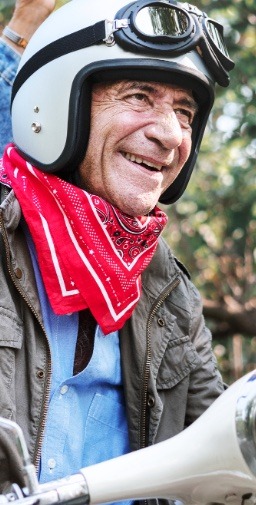 A smiling older man wearing a red bandana and white helmet riding a vespa