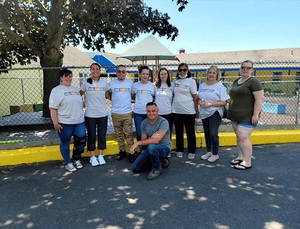 9 smiling people in white ONE Chelsea t-shirts outside of a playground