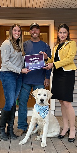A lending banker and a man and woman with their dog on a front porch hold a purple sign with white text that reads "Another Dream Realized! Chelsea Groton"