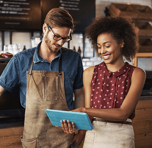 A man in a blue shirt and brown apron and a woman in a red polka dot blouse smile down at a blue tablet inside a cafe