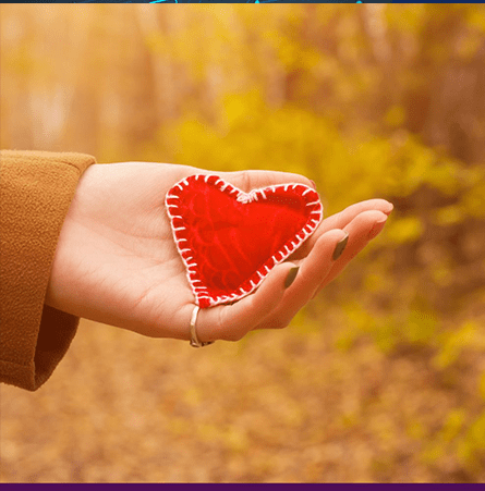 A women's hand with green nail polish and silver ring holding a red felt heart outside with a graphic yellow overlay