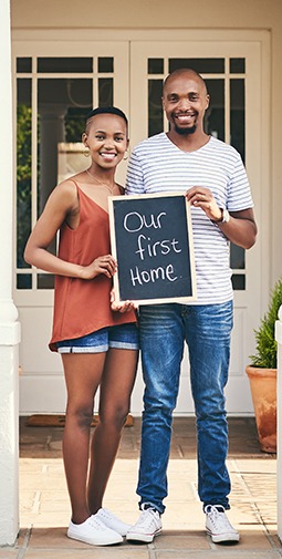 A young couple stands outside a house and holds a black chalkboard with white handwritten text that reads "our first home"