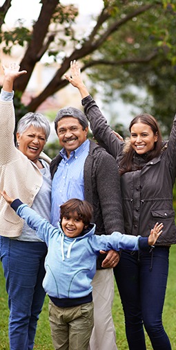 An smiling elderly couple, a mother, and a young child wearing shades of blue and grey raising their arms to the sky in a park 