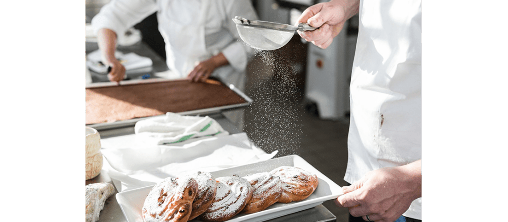 A pastry chef in a kitchen sifting powdered sugar onto pastries