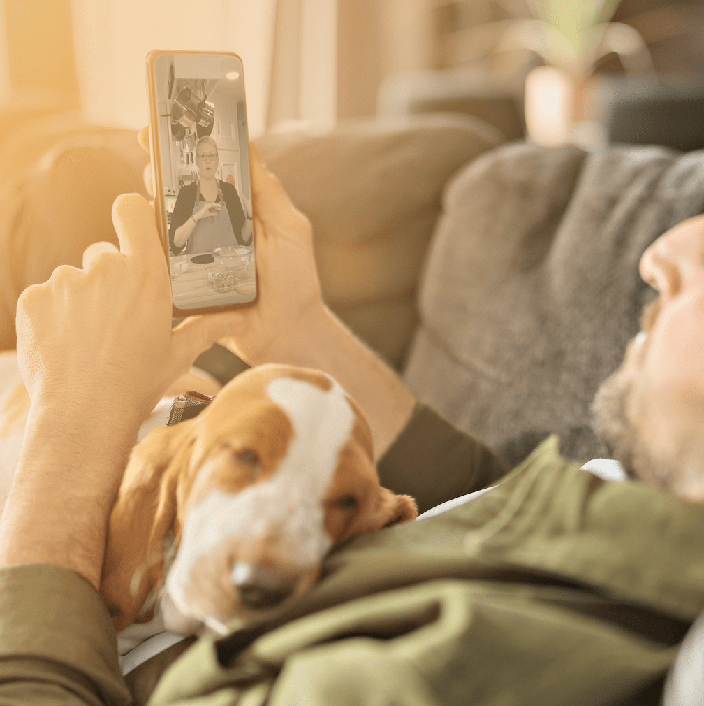 A man laying down on a coach with a dog on his chest watches a cooking video on a smart phone
