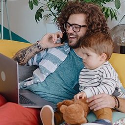 A man with long curly hair on the phone, with his laptop open and his kid in his arm as he sits on the couch